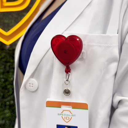 Red heart-shaped badge holder on a white lab coat pocket loop attaching an ID badge against a blurred background. Sold by Specialist ID.