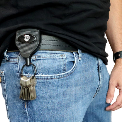 A person wearing blue jeans and a black belt secured with a Customizable Key-Bak Super 48 Heavy Duty Key Reel (S48K) - Customize with Your Own Logo and Design. Several keys are attached to the key holder. The person is also wearing a black shirt and a black wristwatch, showcasing both style and rugged durability.