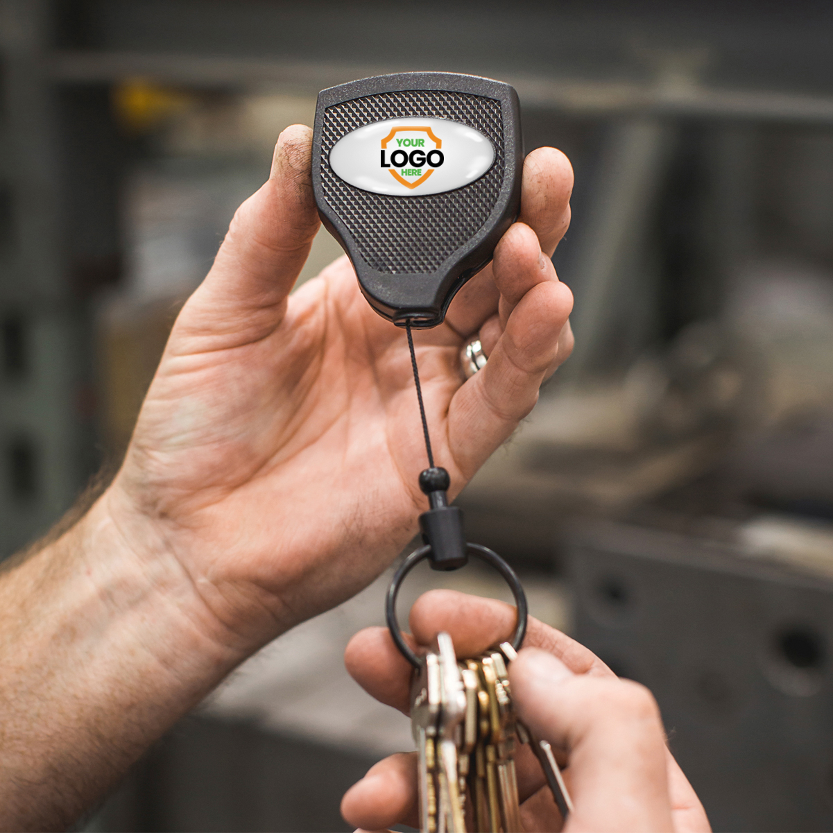 A hand holds a Customizable Key-Bak Super 48 Heavy Duty Key Reel (S48K) - Customize with Your Own Logo and Design with keys attached, displaying a customizable area for a logo. Another hand pulls on the keys, extending the reel's cord with rugged durability.