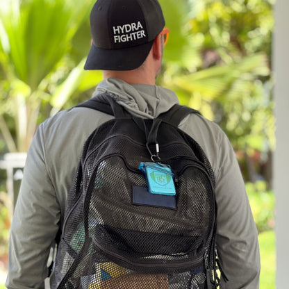 A person in a black "HYDRA FIGHTER" cap and gray hoodie stands outdoors with a black mesh backpack featuring a blue tag and wears the Clear Waterproof ID Badge Holder with Lanyard and Carabiner (P/N SPID-1690).