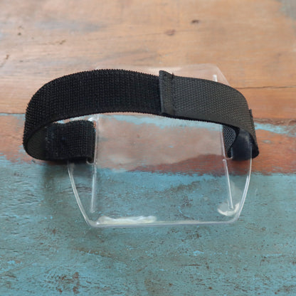 A rectangular clear visor with a black elastic strap, similar to rugged vinyl, sits on a weathered blue and brown wooden surface. The product is Vertical Armband ID Badge Holders with Elastic Band for Smaller Size Arms - Hook and Loop Closure (SPID-1710).