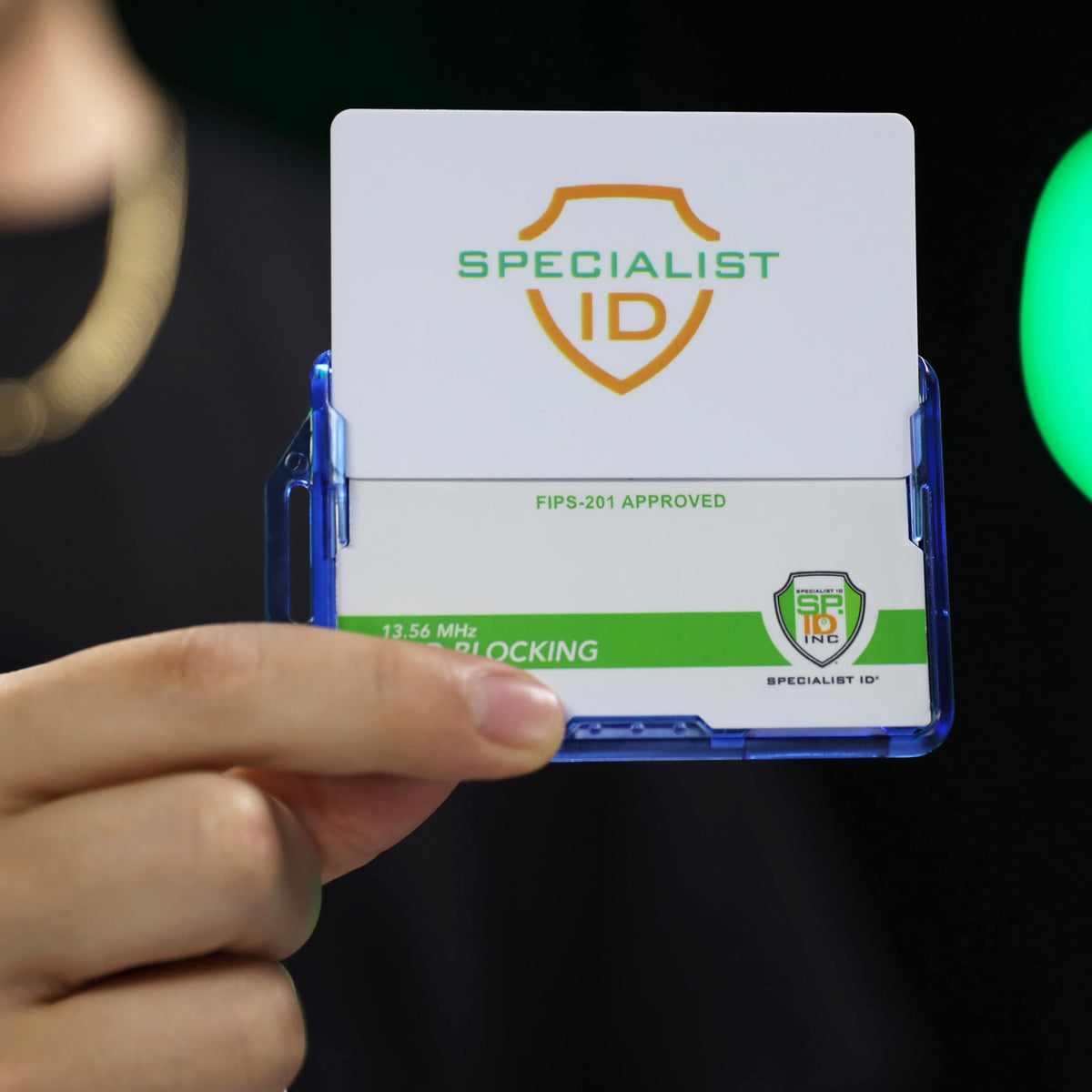 A hand holds a blue Dual RFID Blocking Badge Holder for Smart Cards, CAC, PIV & TWIC (SPID-1720), displaying a white and green security card labeled “SPECIALIST ID” with a shield logo.