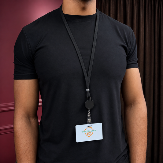 A person wearing a plain black t-shirt and a Breakaway Badge Lanyard with Retractable Reel (SPID-210X) with a "Specialist ID" badge holder stands indoors before dark curtains and a maroon wall.
