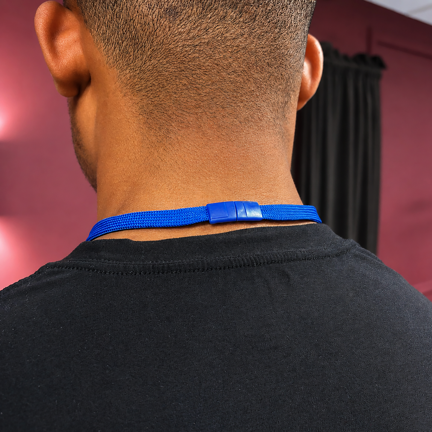 A person in a black shirt wears a Breakaway Badge Lanyard with Retractable Reel (SPID-210X) and ID badge holder around their neck, photographed from behind indoors with maroon walls and a black curtain.