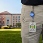 Person wearing beige pants with an ID badge attached to a Custom Printed Oval Shaped Carabiner Badge Reel - Online Designer - Add Personalized Logo or Graphic, standing in front of a building on a grassy lawn.