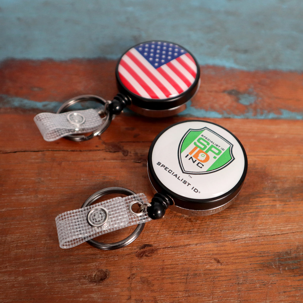Two Custom Heavy Duty Badge Reels with Key Ring and Badge Strap (SPID-3180) - Add Your Logo rest on a wooden surface. One features a US flag design, while the other showcases the "SPECIALIST ID INC" logo. Each reel includes a key ring and badge strap for enhanced functionality and provides a personalized identification display in style.