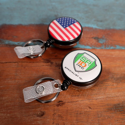 Two Custom Heavy Duty Badge Reels with Key Ring and Badge Strap (SPID-3180) - Add Your Logo rest on a wooden surface. One features a US flag design, while the other showcases the "SPECIALIST ID INC" logo. Each reel includes a key ring and badge strap for enhanced functionality and provides a personalized identification display in style.