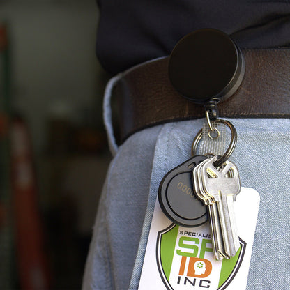 A Heavy Duty Badge Reel with Key Ring and Badge Strap - All Metal Retractor with Belt Clip - Black & Chrome (SPID-3180) is clipped to a person's belt, securely holding a key ring with several keys and an identification badge.