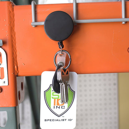 A Heavy Duty Badge Reel with Key Ring and Badge Strap - All Metal Retractor with Belt Clip - Black & Chrome (SPID-3180) is attached to an orange metal surface. The holder has a sturdy belt clip and holds several keys along with a white ID card featuring a green and orange shield design and the text "SPECIALIST ID INC.