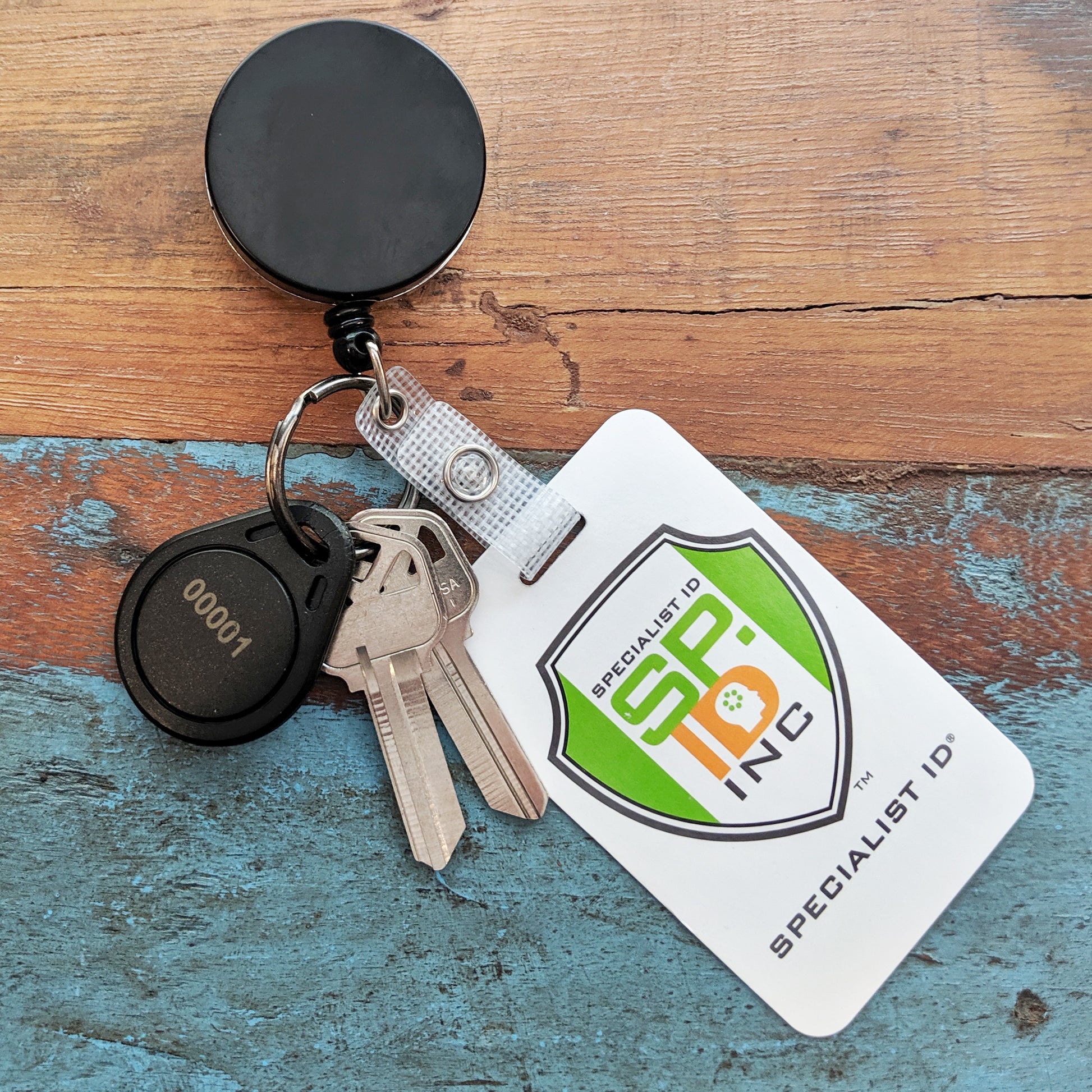 A Heavy Duty Badge Reel with Key Ring and Badge Strap (SPID-3180) holds two keys, a black "00001" key fob, and a Specialist ID badge, all resting on a wooden surface.