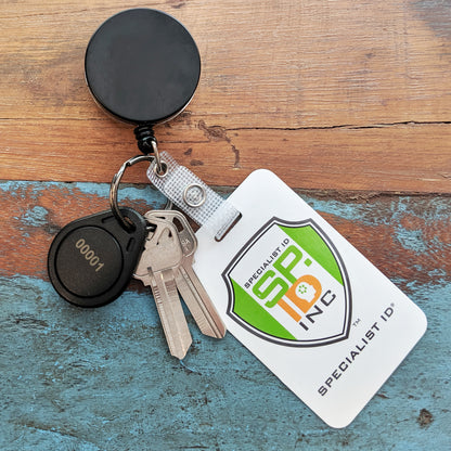 A Heavy Duty Badge Reel with Key Ring and Badge Strap (SPID-3180) holds two keys, a black "00001" key fob, and a Specialist ID badge, all resting on a wooden surface.