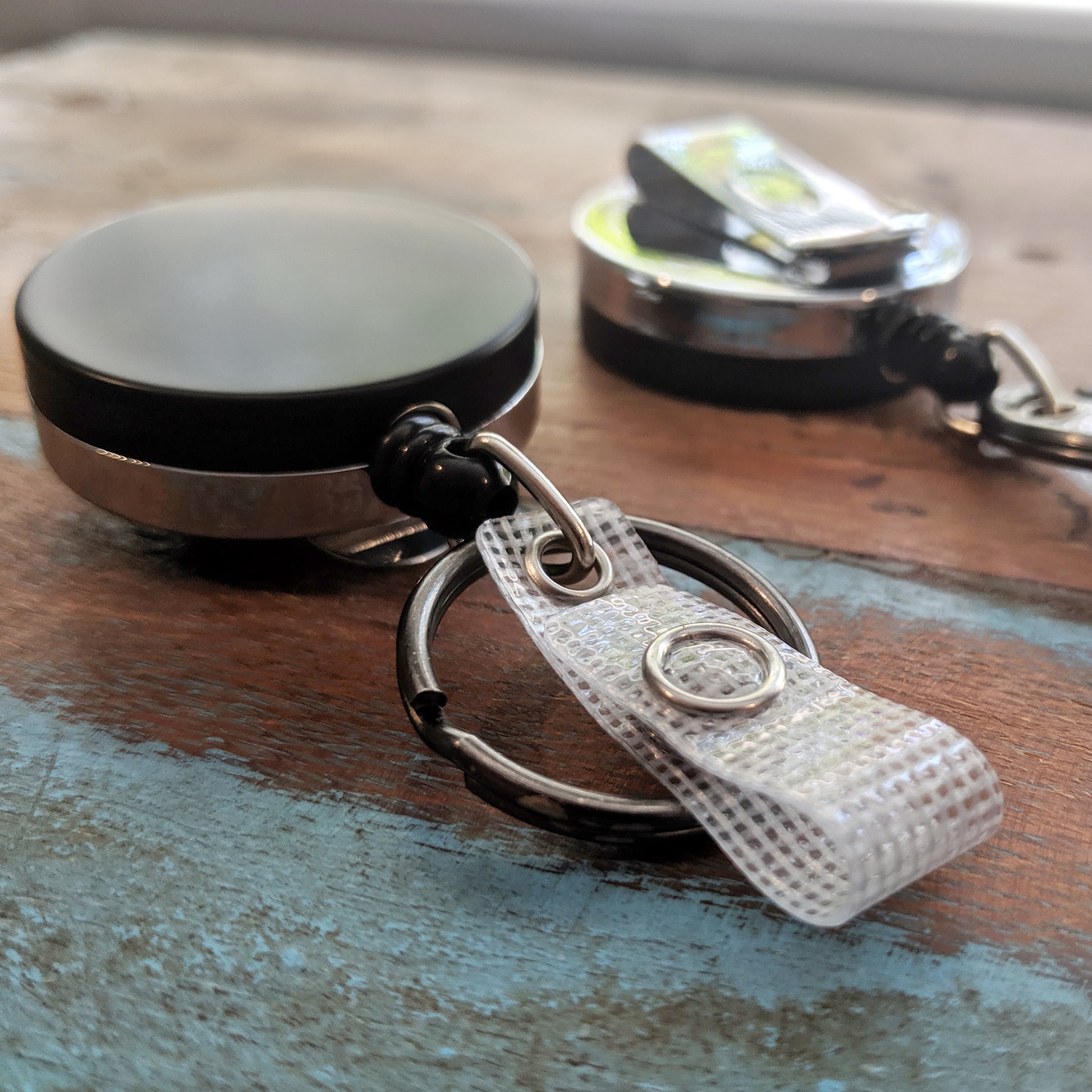 Close-up of the Heavy Duty Badge Reel with Key Ring and Badge Strap - All Metal Retractor with Belt Clip - Black & Chrome (SPID-3180) on wood, another badge reel blurred in the background.