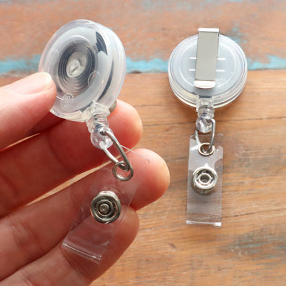 Close-up of a hand holding the Translucent Clear Retractable Badge Reel with Belt Clip (2120-360X) next to another showing the attachment mechanism. Sold by Specialist ID.