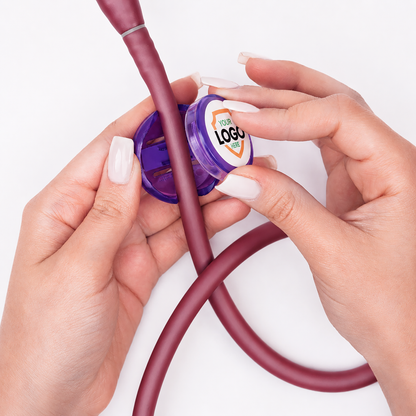Hands attach a circular purple Custom Stethoscope ID Tag, featuring an uploaded logo, to the tubing of a maroon stethoscope on a white background.