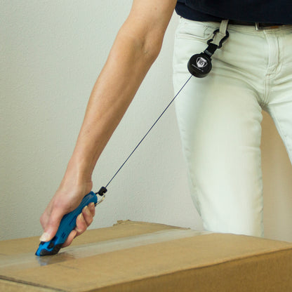 Person using a blue retractable utility knife attached to their belt with a Heavy Duty SPID Key-Bak Mid-Size Carabiner Retractable Badge Reel with Card Strap & Key Ring (SPID-3330) featuring a Kevlar cord to cut tape on a cardboard box.