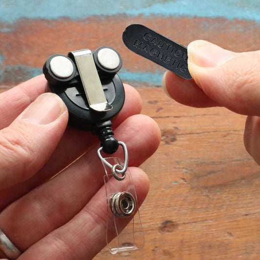 Hands holding a small Magnet Badge Reel - Retractable Magnetic Badge Holder for Shirt Pocket (P/N SPID-3370) with a clip and oval back meta piece that reads 'Caution Magnetic' on a wooden surface.