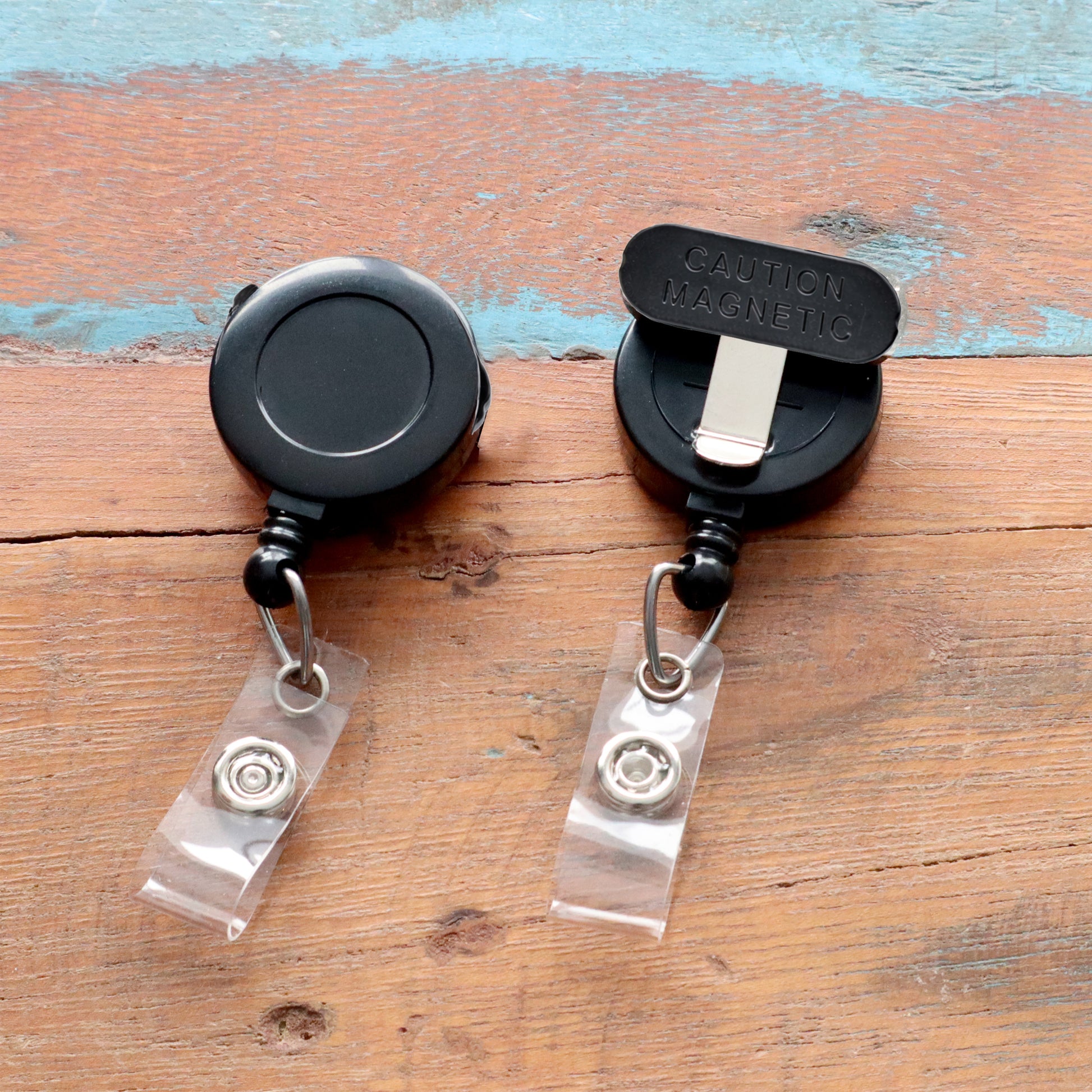 Two Magnet Badge Reels for shirt pockets, P/N SPID-3370, rest on a wooden surface. Their sleek design and retractable feature make them versatile and functional for any name badge setup, blending style with everyday use.