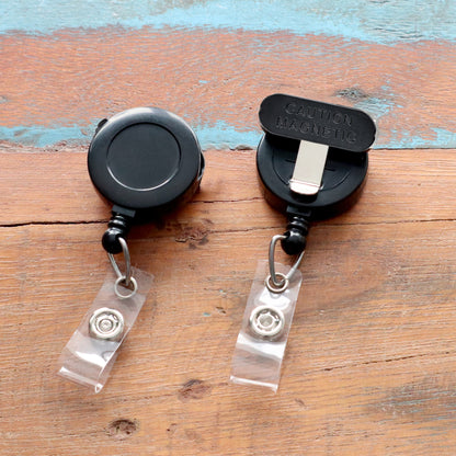 Two Magnet Badge Reels for shirt pockets, P/N SPID-3370, rest on a wooden surface. Their sleek design and retractable feature make them versatile and functional for any name badge setup, blending style with everyday use.