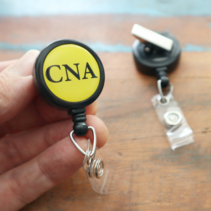 A hand holds the CNA Badge Buddy Reel with Swivel Spring Clip—a yellow retractable reel for nurses—while another badge holder lies on a wooden surface in the background. Sold by Specialist ID.