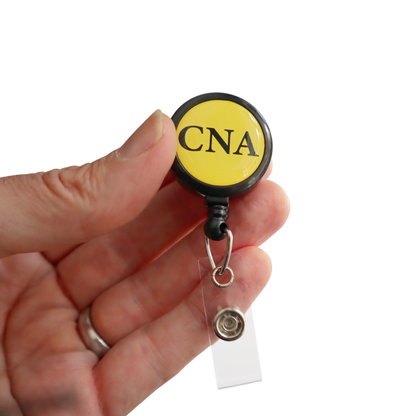 A hand holds the CNA Badge Buddy Reel with Swivel Spring Clip—a round yellow retractable badge holder for nurses, featuring "CNA" in black letters. Sold by Specialist ID.