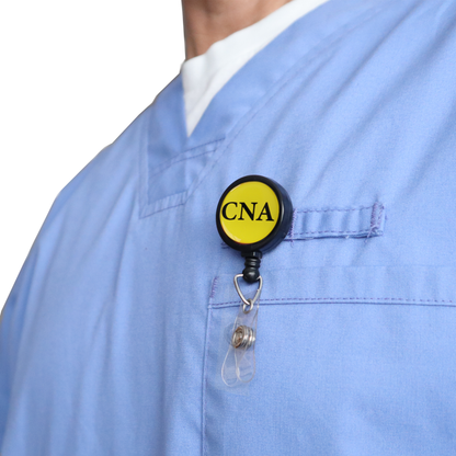 A person wears light blue scrubs with the CNA Badge Buddy Reel with Swivel Spring Clip attached to the chest pocket, combining a yellow "CNA" badge buddy and retractable reel in one for nurses.