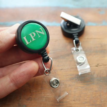 A hand holds the LPN Badge Buddy Reel with Swivel Spring Clip—a green LPN Badge Buddy and retractable reel for nurses—next to a similar unlabeled badge reel on a wooden surface. Sold by Specialist ID.