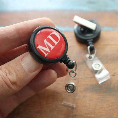A hand holds the MD Badge Buddy Reel with Swivel Spring Clip—an all-in-one retractable reel and badge buddy for doctors; another rests on a wooden surface in the background. Sold by Specialist ID.