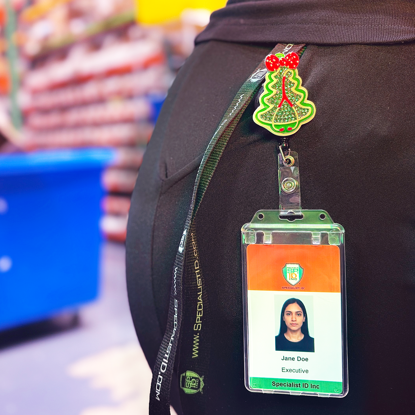 Christmas tree ID badge holder attached to a vertical clear ID badge holder displaying a single ID badge. The Xmas  tree badge reel is clipped ot the side pocket of a person's pants while at work. 
