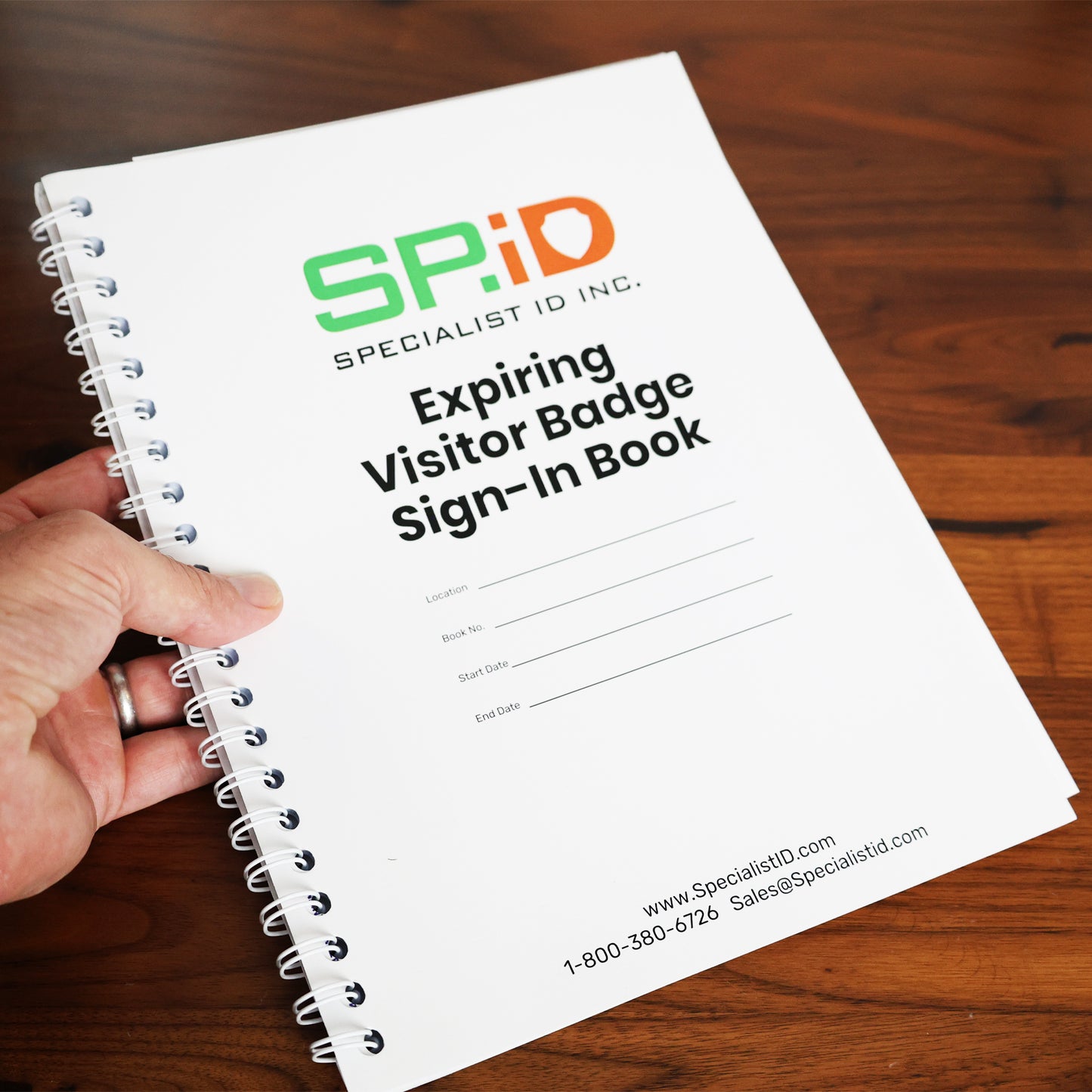 A hand holds the spiral-bound "Expiring Visitor Badge Stickers Log Book – One-Day Security Badges with Sign-In Records," featuring blank fields for location, date, and time on the cover. No SEO keywords are present in this description.