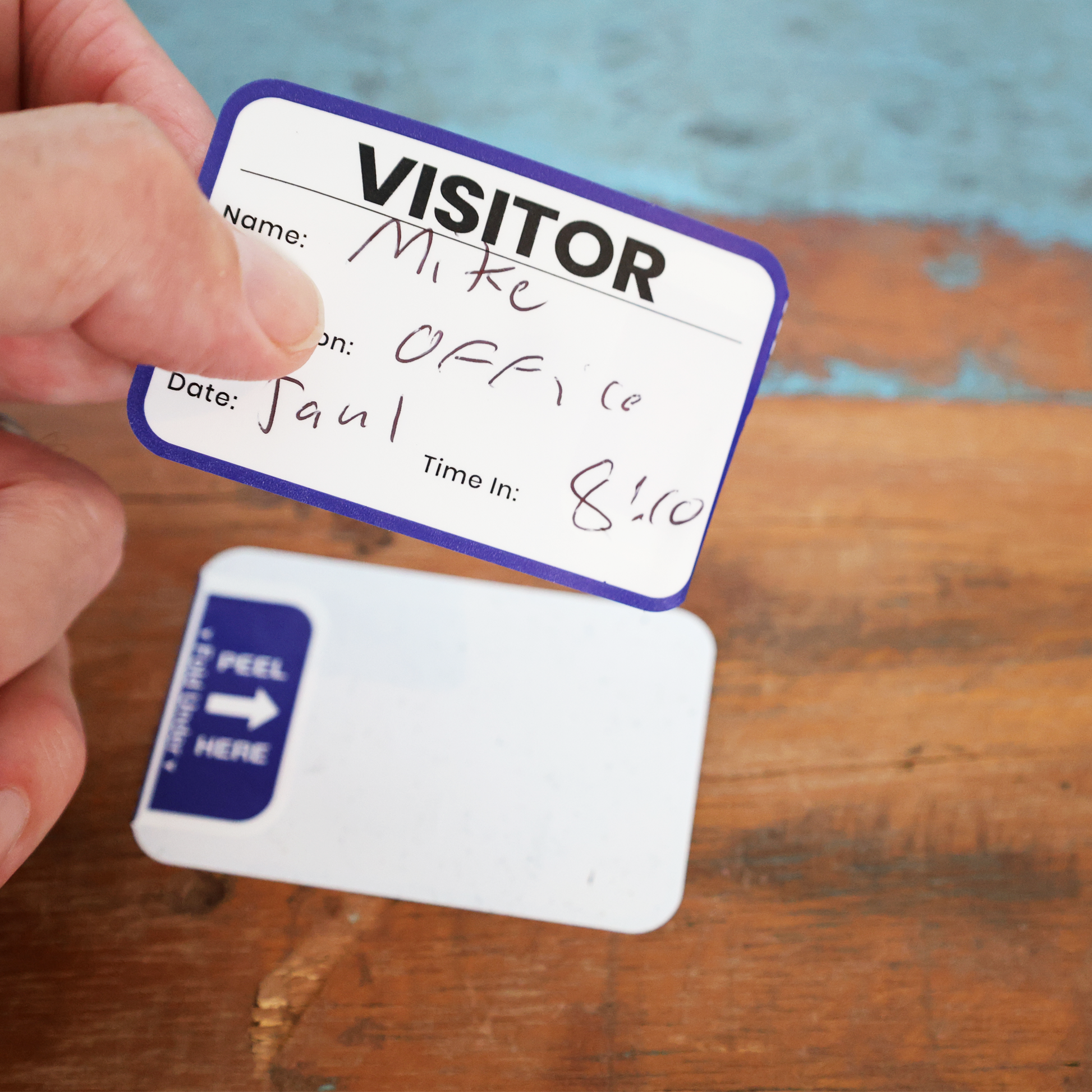 A hand holds an Expiring Visitor Badge Sticker from the Sign-In Log Book, labeled with Mike’s name, office location, Jan 1 date, and 8:10 time in. On the wooden surface below is another sticker marked "peel here.
