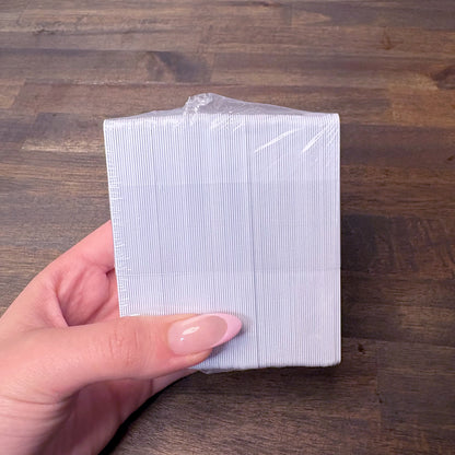 A hand holding a sealed, plastic-wrapped stack of 500 Pack – Professional CR80 30 mil Composite PVC/PET Cards (compatible with most printers) against a wooden surface.