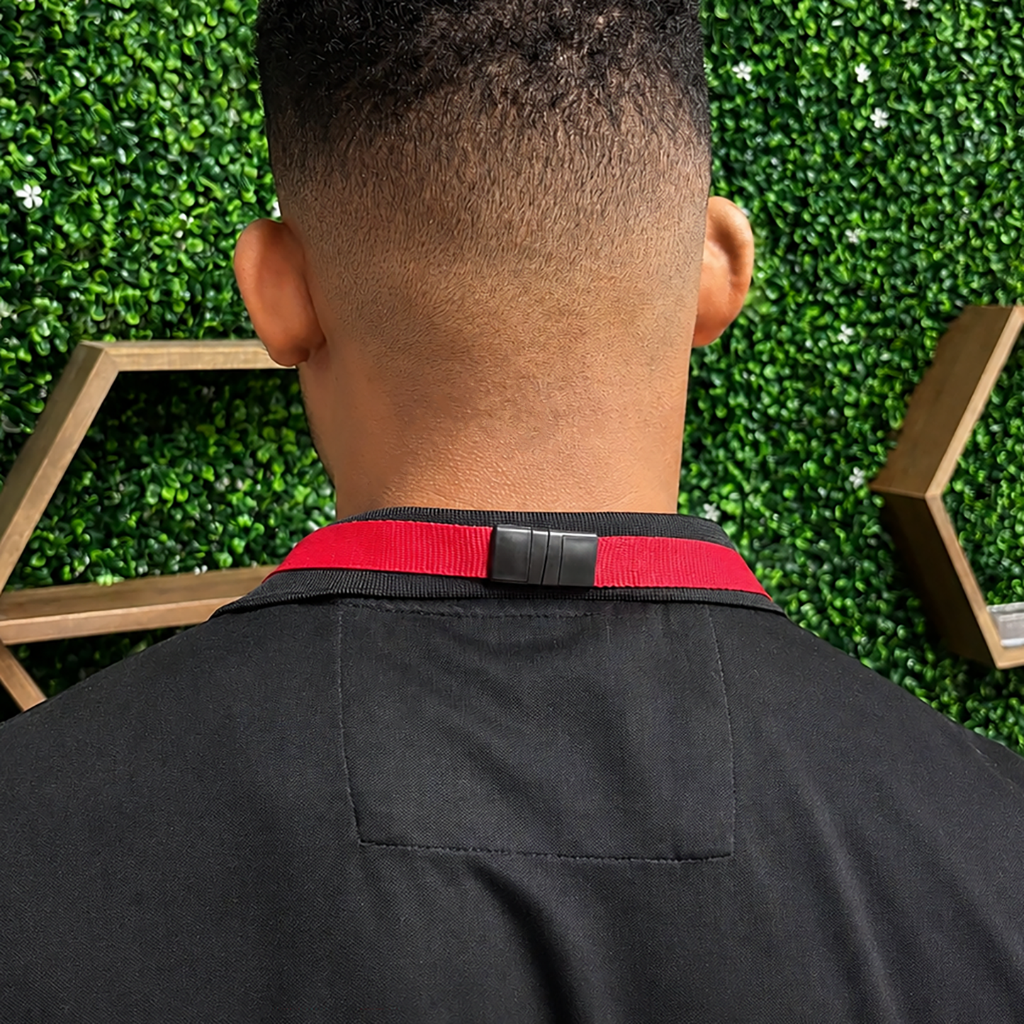 A person with short hair, seen from behind in a black garment with a red collar, wears the Premium Wide Breakaway Lanyard with Detachable Lobster Claw Clasp (2138-362X) while standing before a leafy wall and wooden hexagonal shelves.