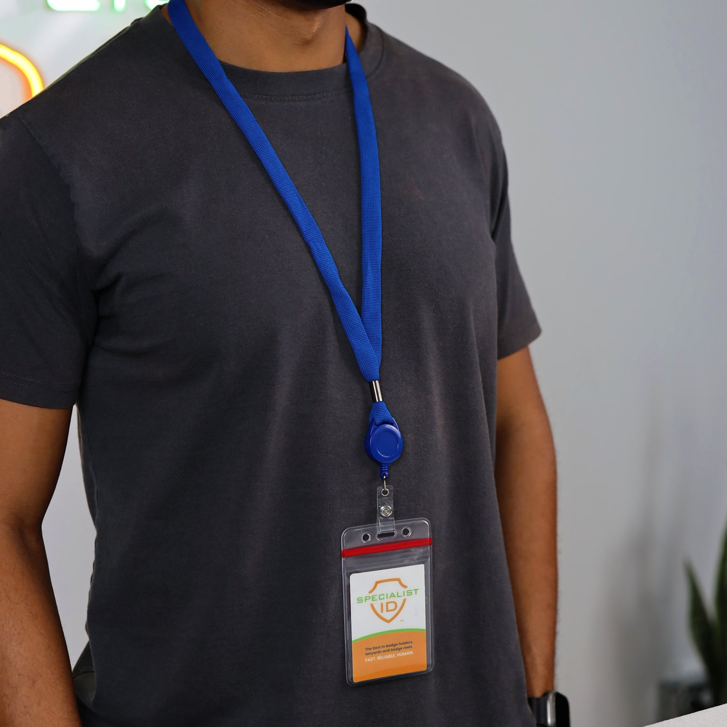 A person wearing a dark t-shirt and a royal blue lanyard displays an ID badge attached to the “Cruisin Through Life™ Retractable Badge Holder” from the 5 Pack Family Pack set. The background is blurred, with a plant visible on the right side.