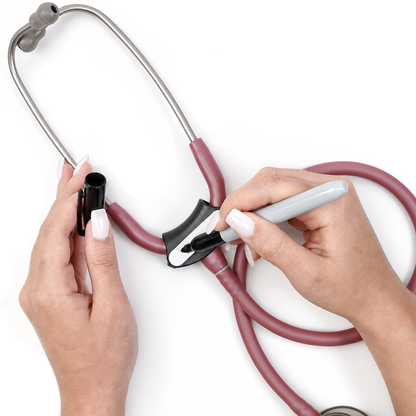 A close-up of a person's hands holding a stethoscope and using a black marker to write on the blank white label of the black stethoscope tag attached to the medical stethoscope tubing, set against a plain white background.