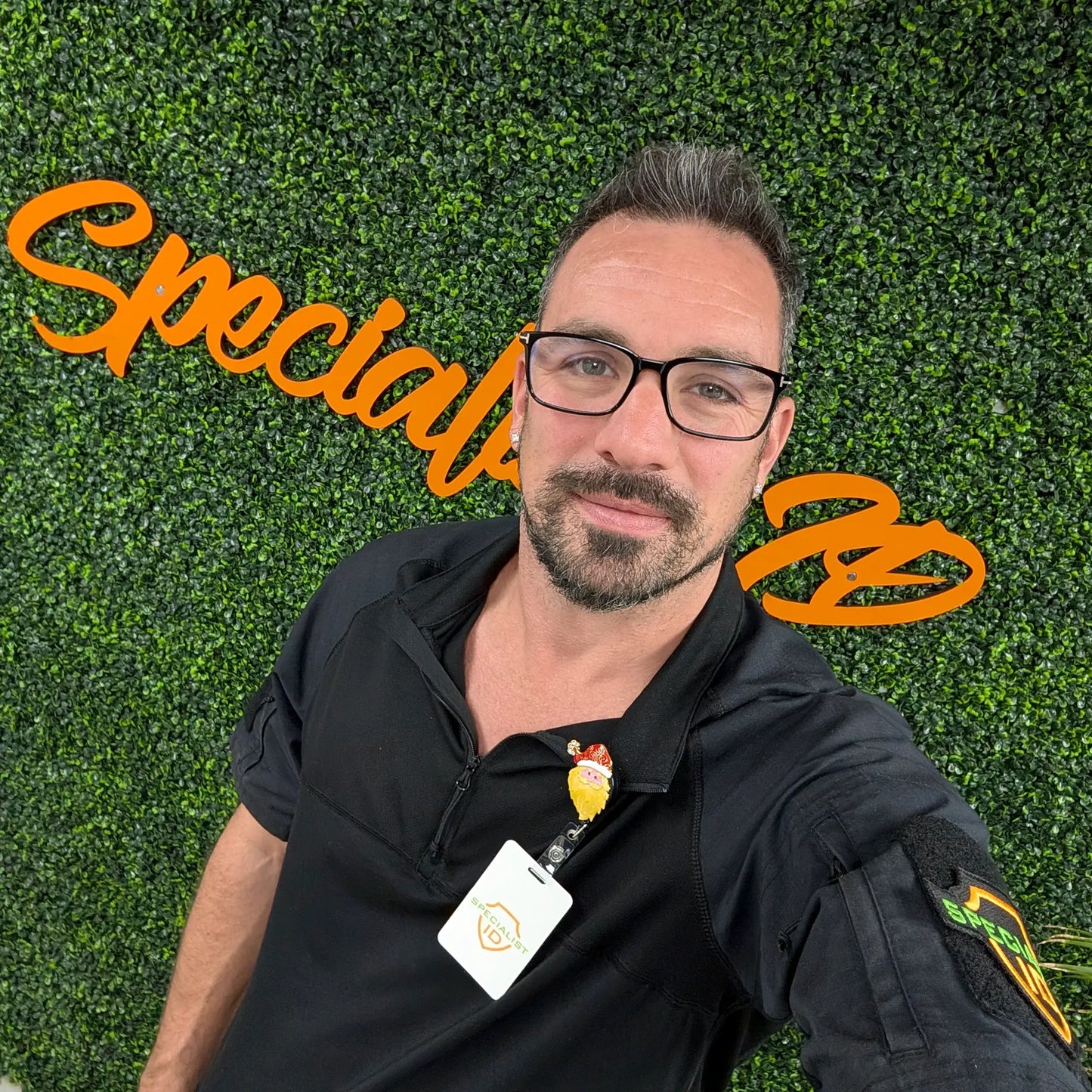 A bearded man with glasses takes a selfie by a green leafy wall with orange "Special" text, wearing a black shirt and an ID badge on the Holiday Santa Claus Badge Reel – Cute Festive ID Holder for Nurses, Staff & Professionals.