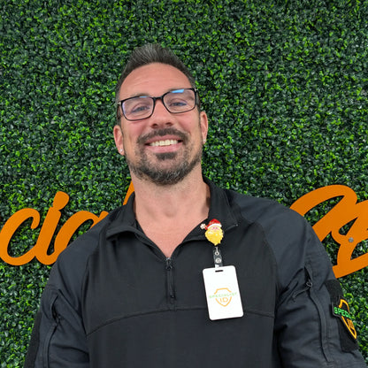 A man in glasses and a black jacket stands before a green leafy wall with orange lettering, smiling at the camera. He has the Holiday Santa Claus Badge Reel – Cute Festive ID Holder for Nurses, Staff & Professionals clipped to his jacket.