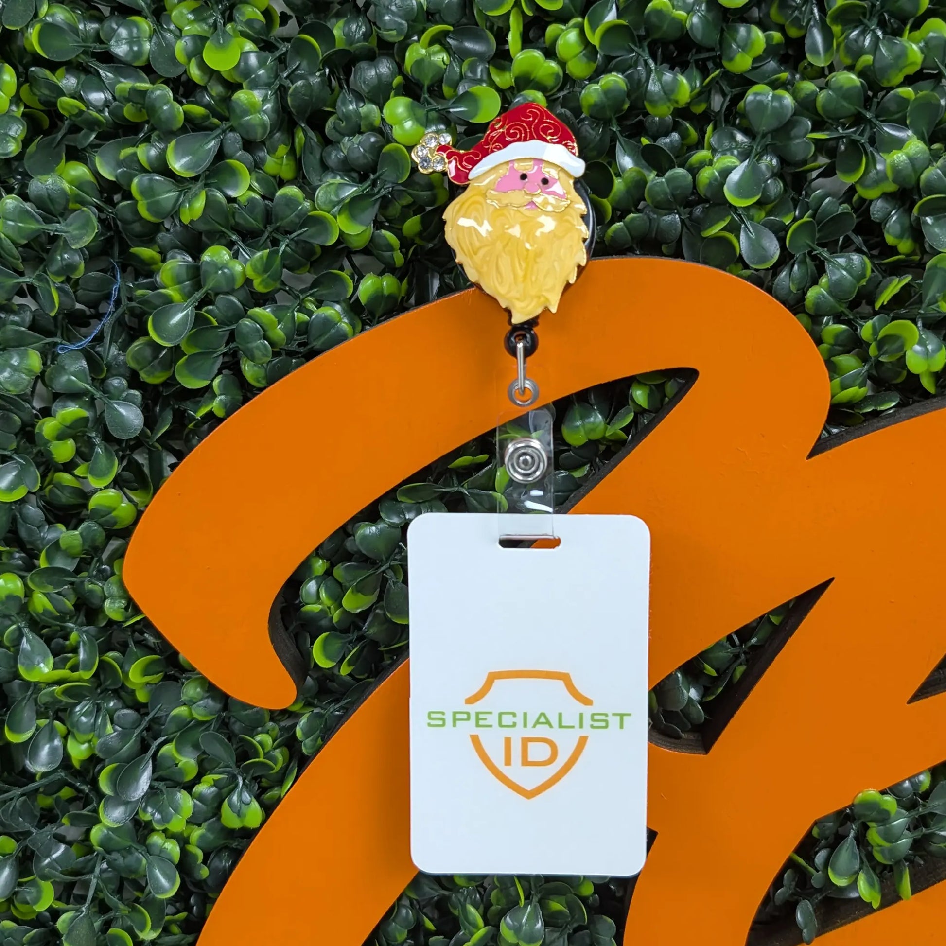 A Specialist ID badge hangs on an orange structure, attached to the Holiday Santa Claus Badge Reel – Cute Festive ID Holder for Nurses, Staff & Professionals, with a green leafy background.