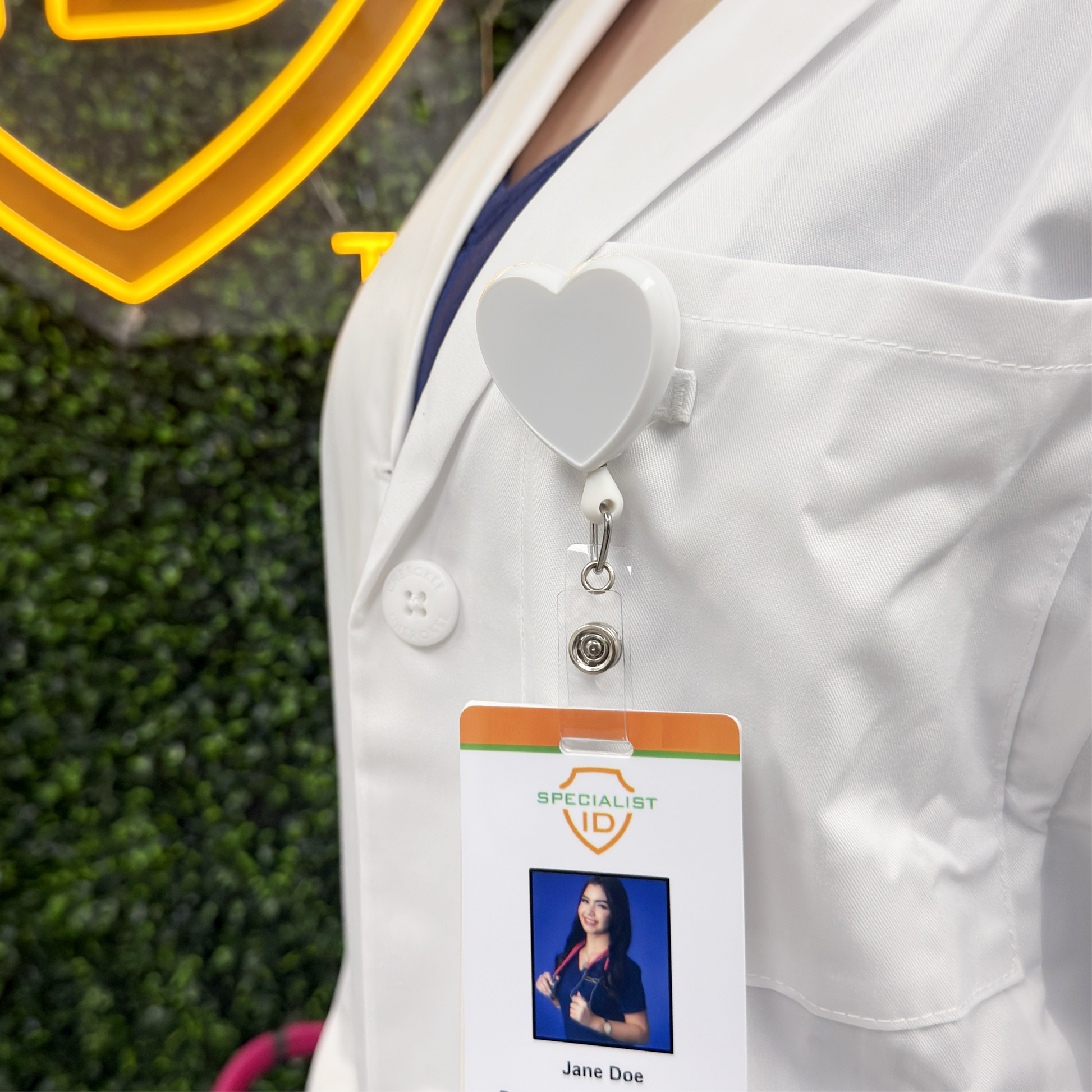 A person in a white lab coat wears a Heart Shaped Badge Reel With Rotating Spring Clip (P/N 2120-761X) displaying their ID, photo, name, and "SPECIALIST ID" label—making it a thoughtful nurse gift.