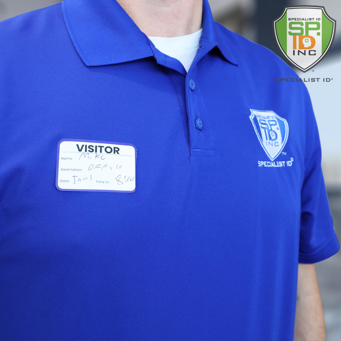 A person wearing a blue polo shirt with the Specialist ID logo and an Expiring Visitor Badge Stickers Log Book one-day security badge labeled “Mike” stands in front of the Specialist ID company logo.