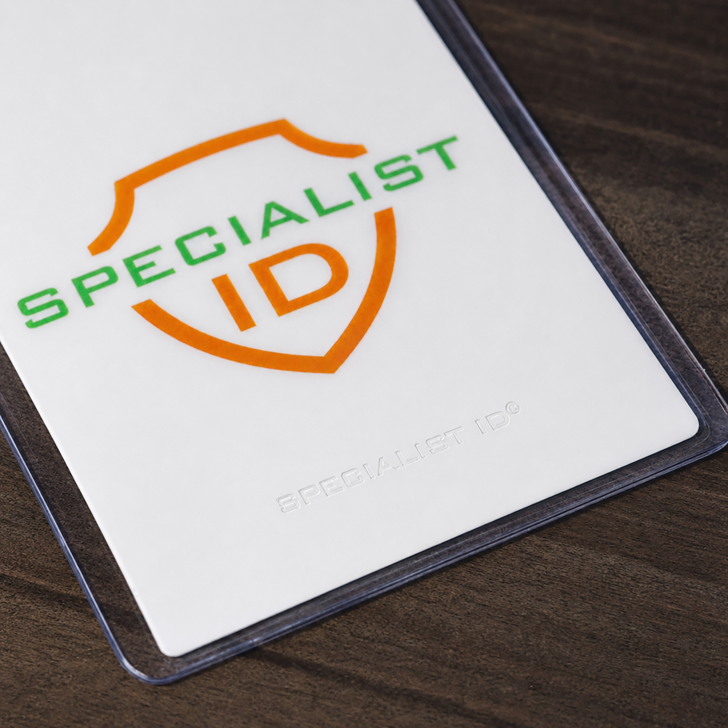 A close-up of a Vertical 4x6 Oversized Vinyl Badge Holder (XL46V) with the words “SPECIALIST ID” in green and orange, displayed inside a clear plastic holder on a dark wood surface.