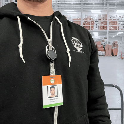 A person in a black hoodie and "Raphael Smith, Operations" badge stands in a warehouse, their ID attached to a Carabiner No Twist Badge Reel (P/N 704-CB) among shelves and orange-wrapped items.