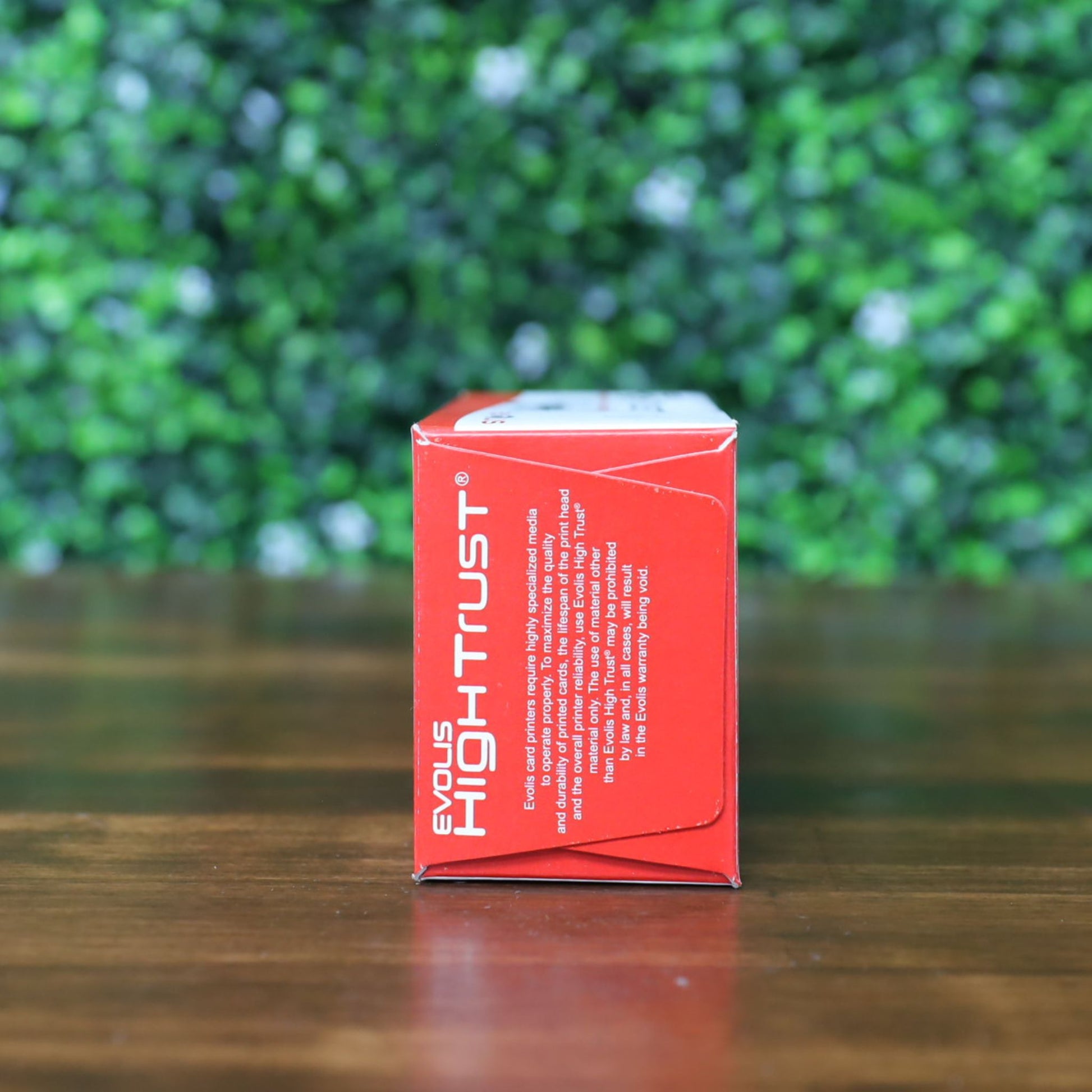 Side view of Evolis High Trust R3314 YMCKOK Ribbon box resting on its side, showing the red end panel with white product and brand text. Displayed on a wooden surface with a blurred green leafy background. Photographed and sold by Specialist ID, an authorized Evolis dealer.