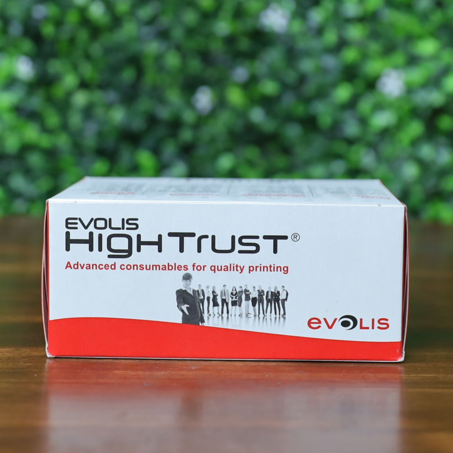 Side view of Evolis High Trust R3314 YMCKOK Ribbon box featuring red-and-white design and silhouette image of professionals. Box is positioned upright on a wooden surface with a blurred green leafy background. Photographed and sold by Specialist ID, an authorized Evolis dealer.