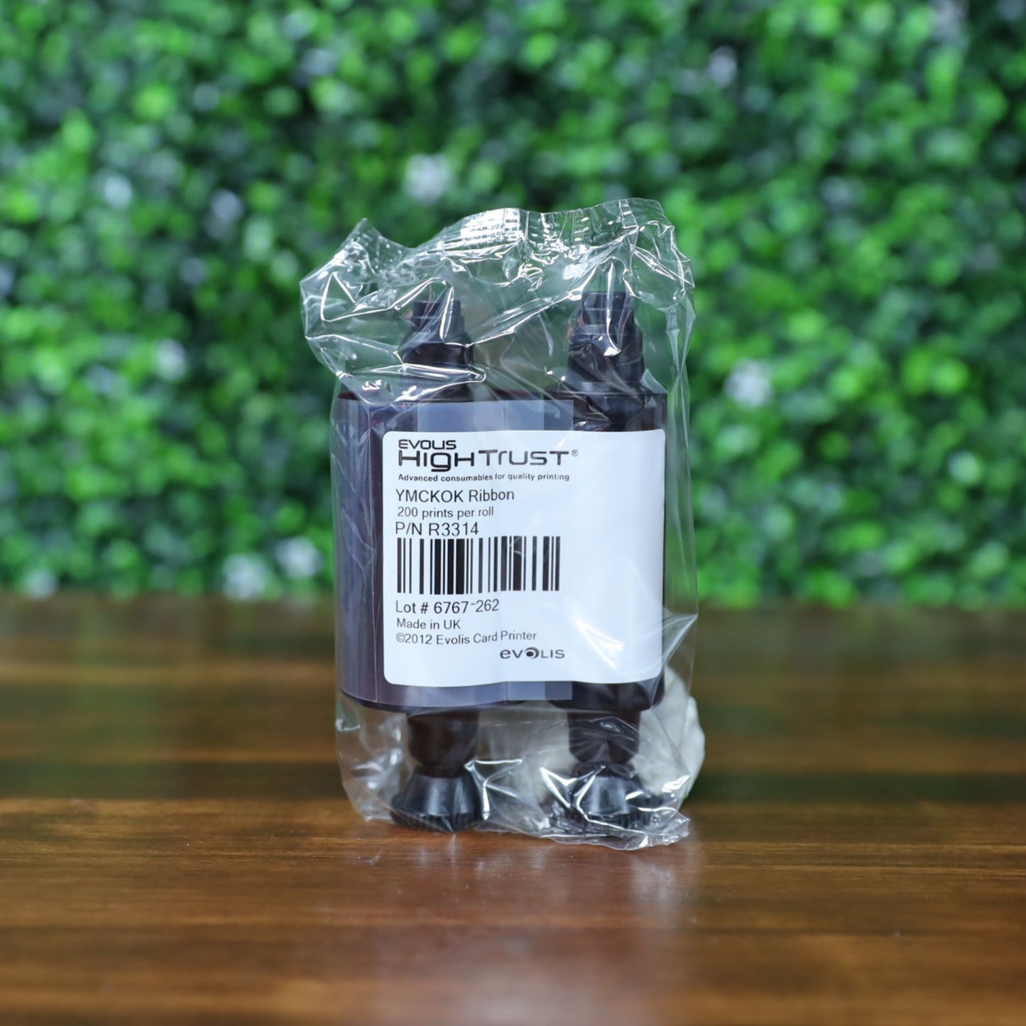 Sealed Evolis R3314 YMCKOK Ribbon – 200 prints, shown inside original twin-spool plastic packaging with barcode label visible, placed on a wooden surface with a leafy green background. Photographed and sold by Specialist ID, an authorized Evolis dealer.