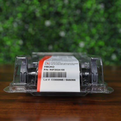 Close-up of sealed Evolis R5F202A100 YMCKO ribbon cartridge in High Trust packaging for Primacy 2 ID Card Printer, showing printed label with part number and lot number on clear plastic case, placed on wood table with green foliage background. Photographed and sold by Specialist ID, an authorized Evolis dealer.
