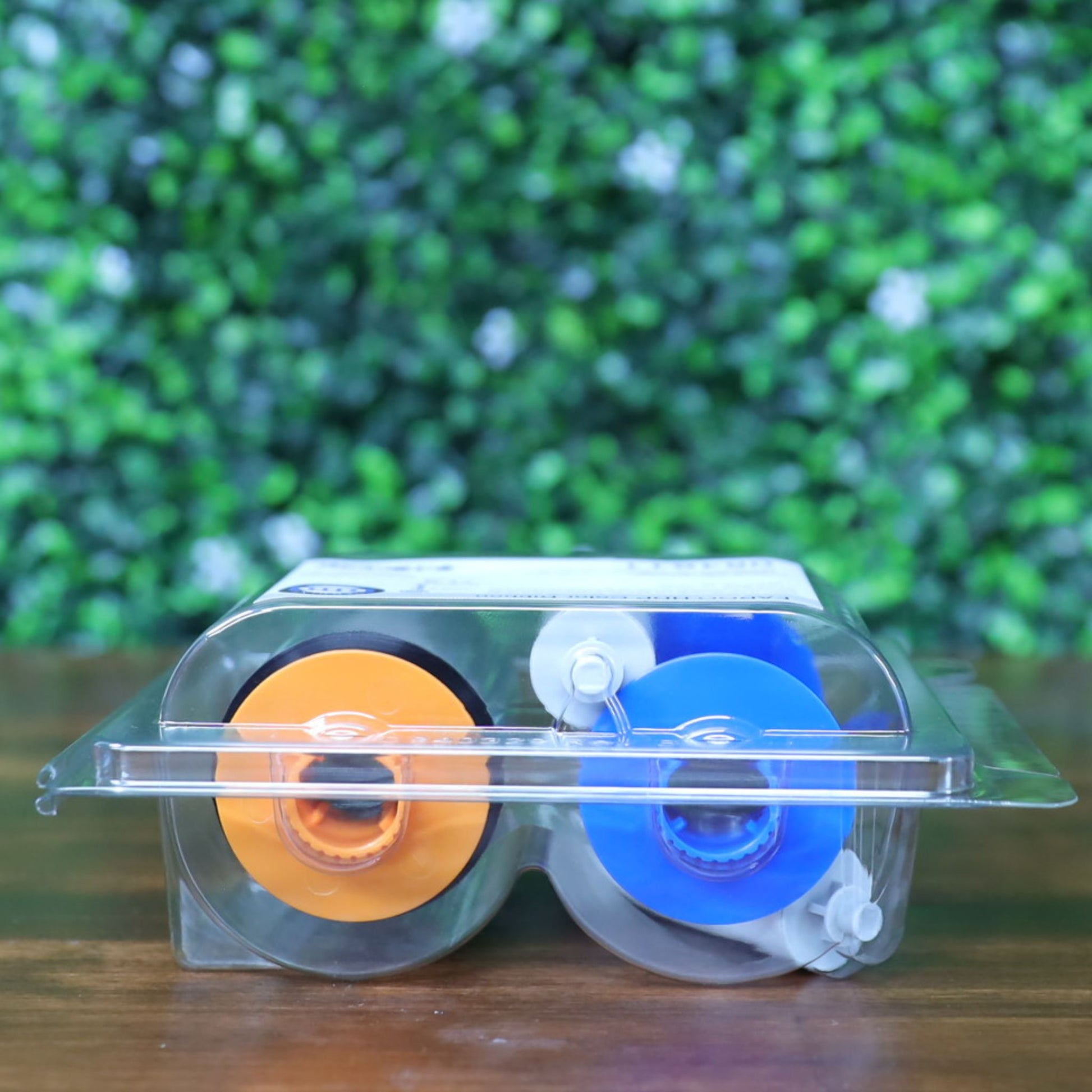 Bottom view of HID Fargo 84811 YMCK color ribbon in clamshell packaging, showcasing bright orange and blue spool cores lying flat on a wood surface with a green blurred background. Photographed and sold by Specialist ID, an authorized HID Fargo dealer.
