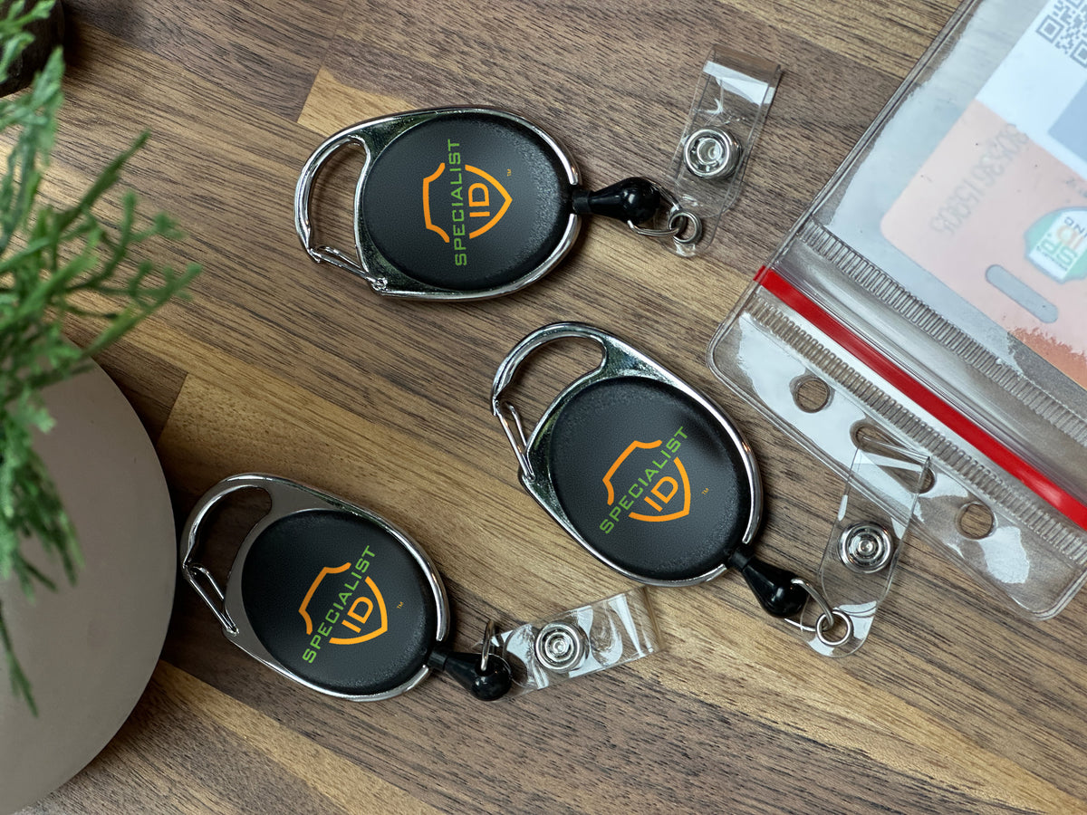 Three Specialist ID branded carabiner badge reels and a clear badge holder on a wooden desk