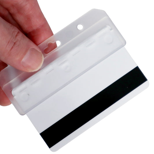A hand holds a blank white plastic card with a black magnetic stripe, placed inside a Swiple Card Holder - Rigid Horizontal Half Card Badge Holder - Leaves Mag Stripe Exposed for Easy Swiping (1840-8000) with holes for attachment.