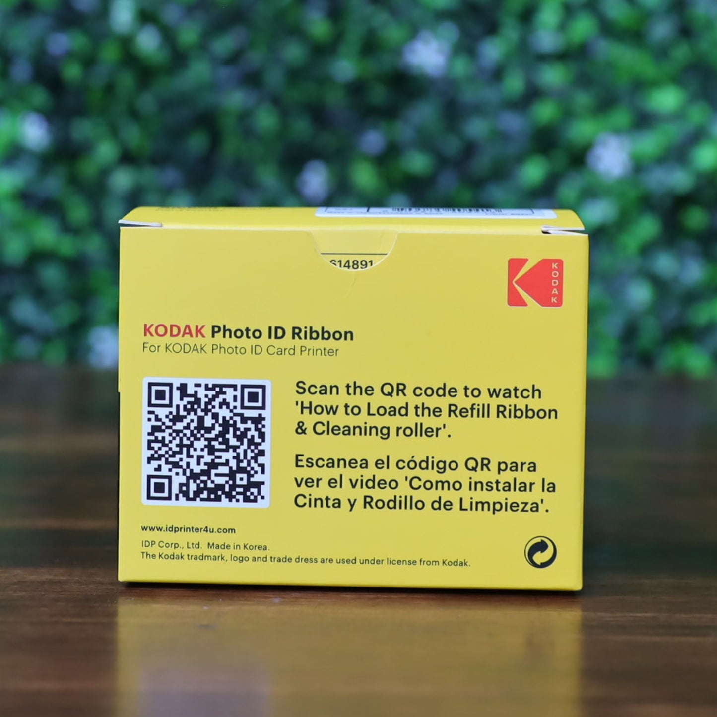 KODAK 653750 Photo ID Color Ribbon for KODAK ID100S and ID200S card printers (YMCKO, 100 prints) in a yellow box with QR code and bilingual instructions for loading the ribbon and cleaning roller, displayed on a wooden surface with a blurred green leafy background. Photographed and sold by Specialist ID, an authorized Magicard dealer.