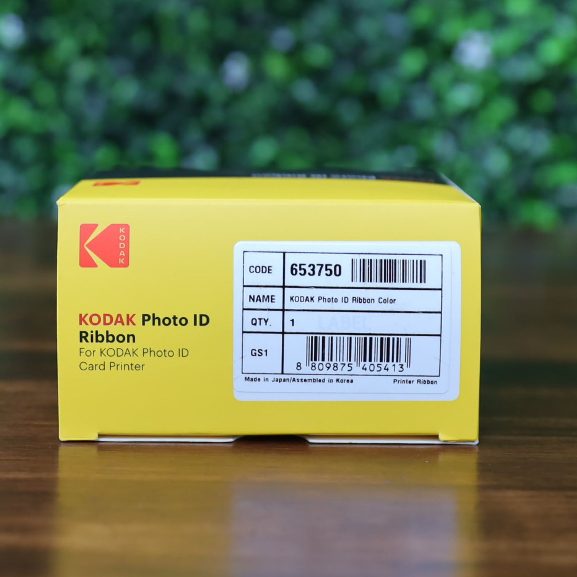 A KODAK 653750 Photo ID Color Ribbon YMCKO (100 prints) for KODAK ID100S and ID200S card printers in its yellow box with barcode label, displayed on a wooden surface with a blurred green leafy background. Photographed and sold by Specialist ID, an authorized Magicard dealer.